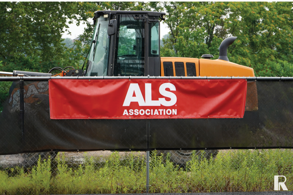 Red banner with the text “ALS ASSOCIATION” attached to a chain-link fence in front of construction equipment.