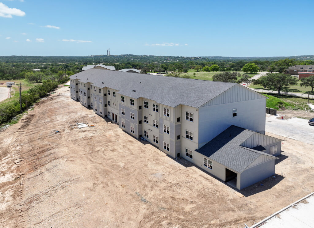 Aerial view of a multi-story apartment building under construction, surrounded by dirt ground and a rural landscape.