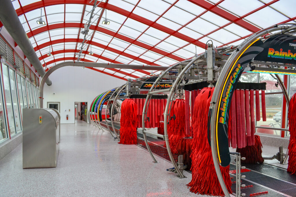 The interior of a car wash with a curved glass roof, featuring multiple red spinning brushes and equipment labeled “RainbowCoat.”