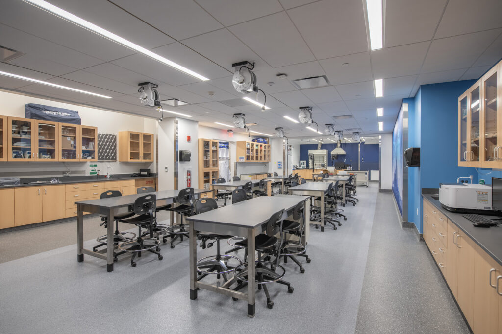 A science laboratory classroom with rows of metal tables, black rolling chairs, light wood cabinets, and overhead retractable power reels.