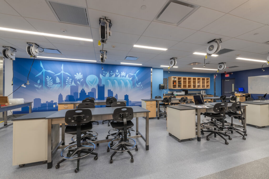 A modern science classroom featuring modular desks, swivel chairs, and a large wall mural depicting a city skyline and sustainable energy symbols.