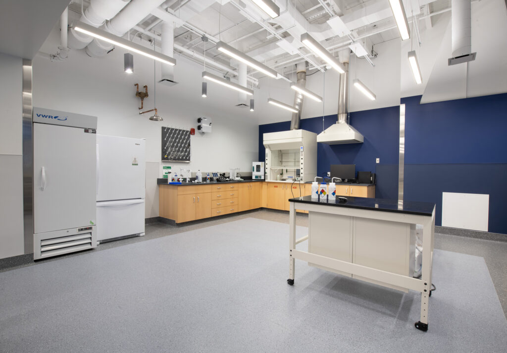 A laboratory with light wood cabinets, a fume hood, a tall white refrigerator, and a mobile workstation.