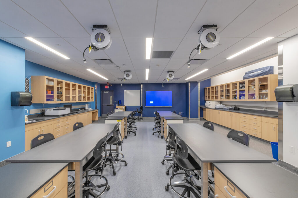 A science laboratory classroom with two rows of gray tables, stools, light-wood cabinets, and retractable power reels hanging from the ceiling.