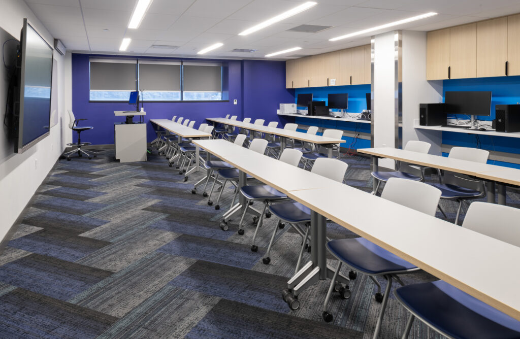 Modern computer classroom with rows of desks and chairs facing a wall-mounted display and blue accent walls.