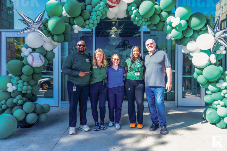 Five employees stand together in front of a storefront entrance decorated with a large arch of green and white balloons featuring a baseball graphic.