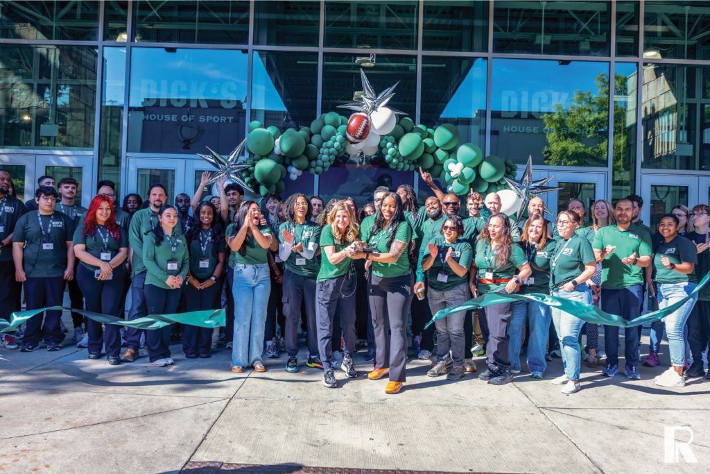 A large group of people in green Dick’s House of Sport shirts celebrating with a ribbon-cutting ceremony in front of a store entrance decorated with green balloons and a football.