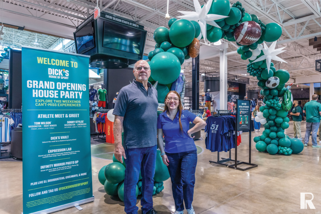 A man and a woman stand in a sporting goods store in front of a green balloon arch and a grand opening event sign for Dick’s House of Sport.