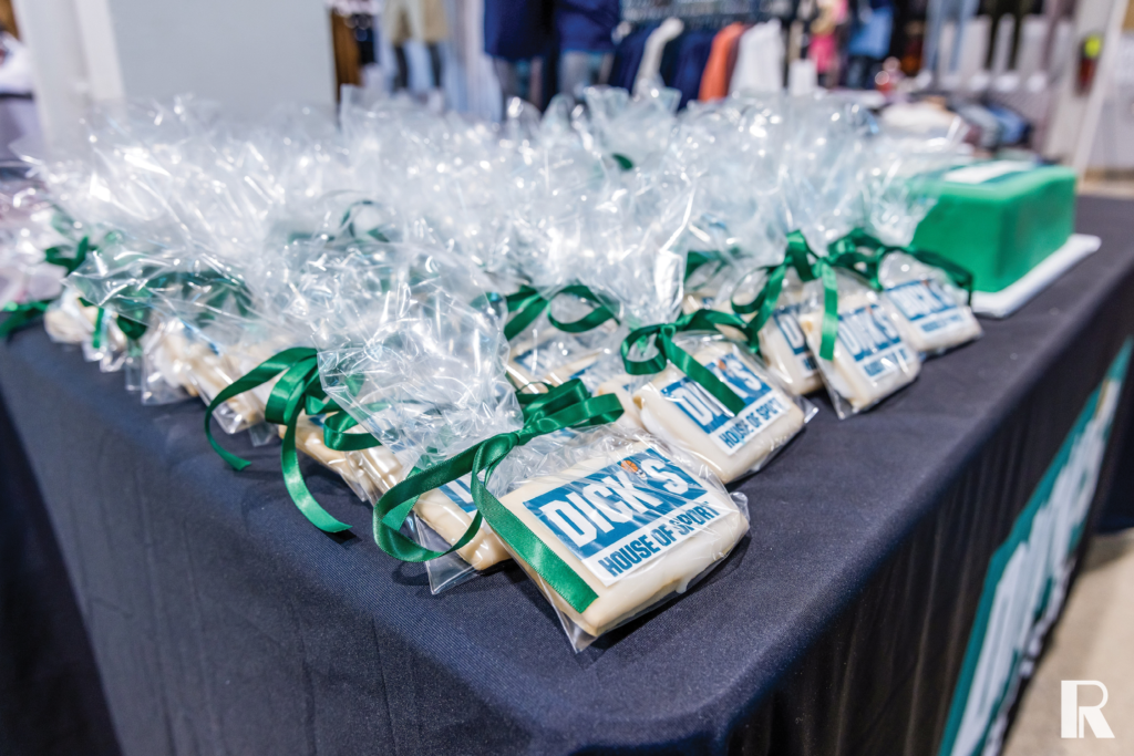Dozens of square sugar cookies with the Dick’s House of Sport logo, individually wrapped in clear plastic bags with green ribbons on a black table.