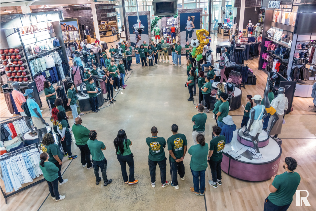 A group of employees in green shirts stands in a circle inside a retail store during a meeting.