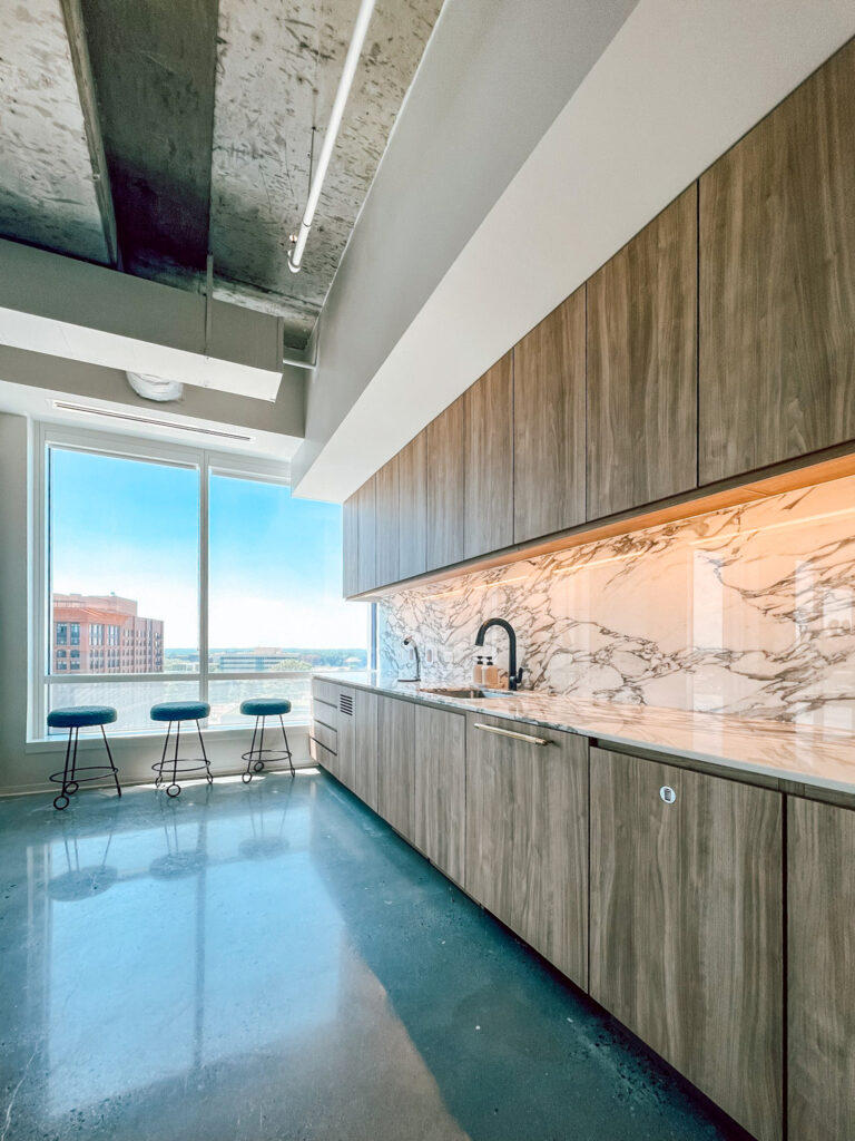 Modern kitchen with wood cabinetry, marble backsplash, and a row of three stools by a large window overlooking a city skyline.