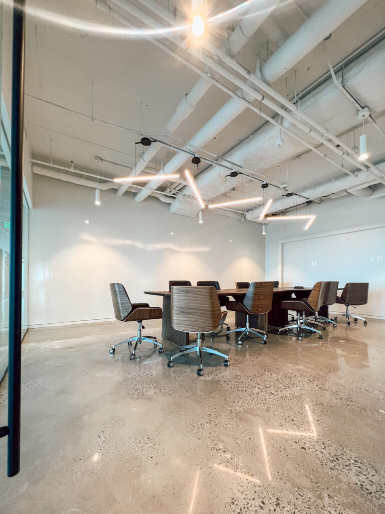 A conference room with a long table surrounded by modern wood-finish chairs under exposed ceiling pipes and linear light fixtures.