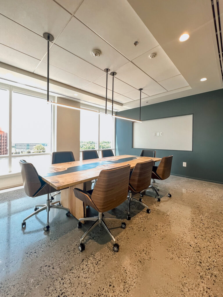 A modern conference room featuring a live-edge wooden table with a black resin inlay, surrounded by six wheeled office chairs under minimalist pendant lights.
