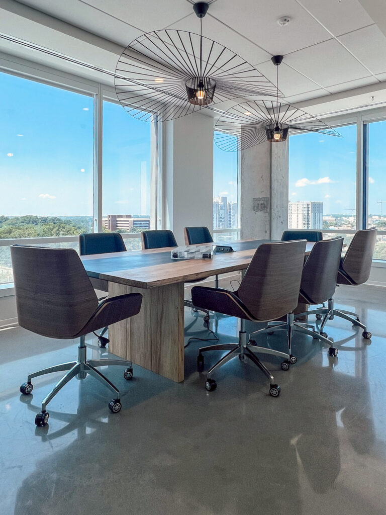 A conference room with a long wood table and office chairs, positioned beneath two large, circular wire pendant lights in front of floor-to-ceiling windows.