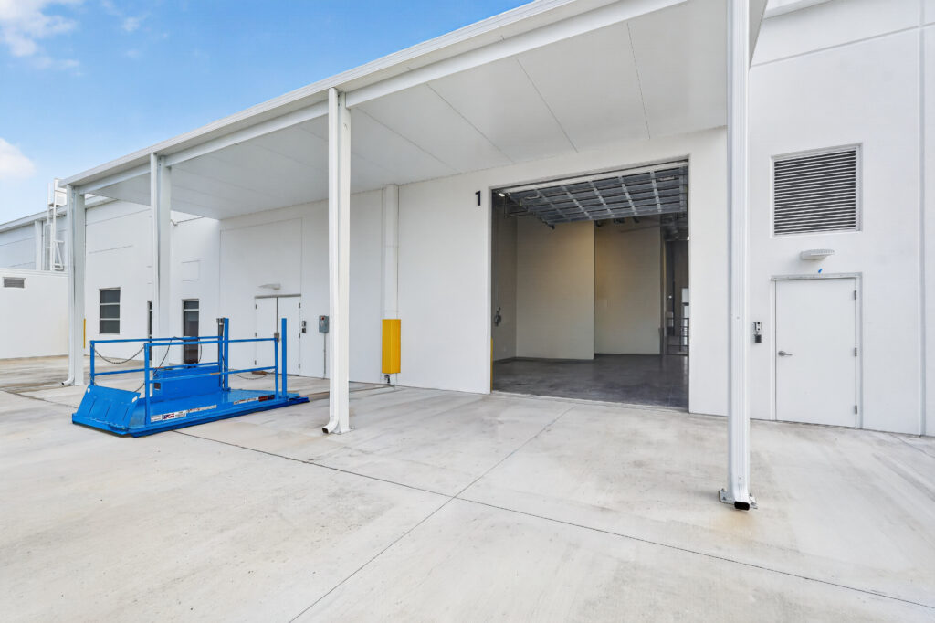 Exterior view of a commercial building with a loading dock, an open bay door marked with the number 1, and a blue portable lift platform.