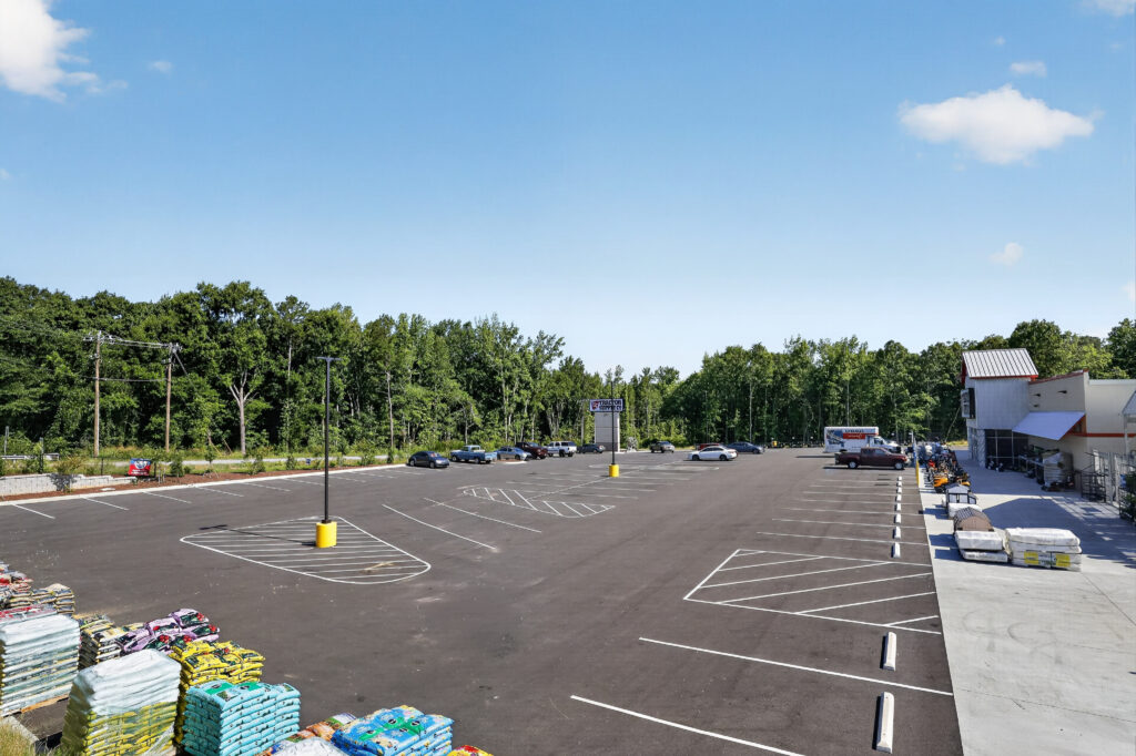 Empty parking lot in front of a Tractor Supply Co. store, with stacks of bagged goods in the foreground and a line of trees in the background.