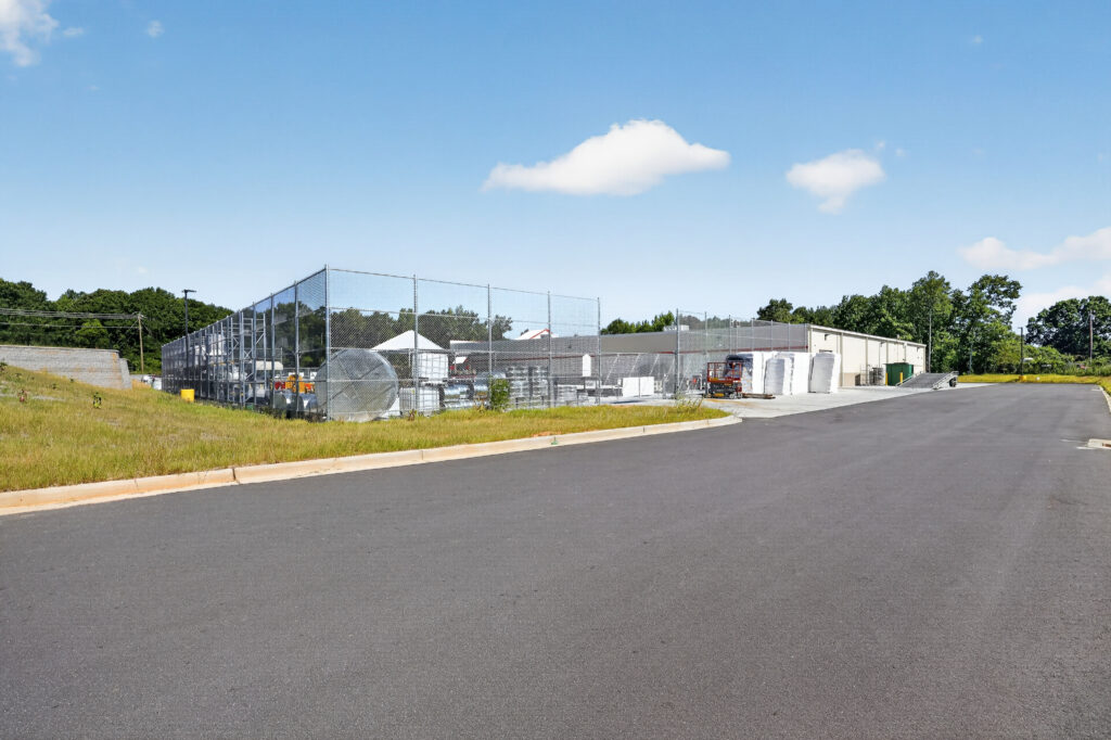A chain-link fenced storage yard behind a commercial building, adjacent to an asphalt parking lot.