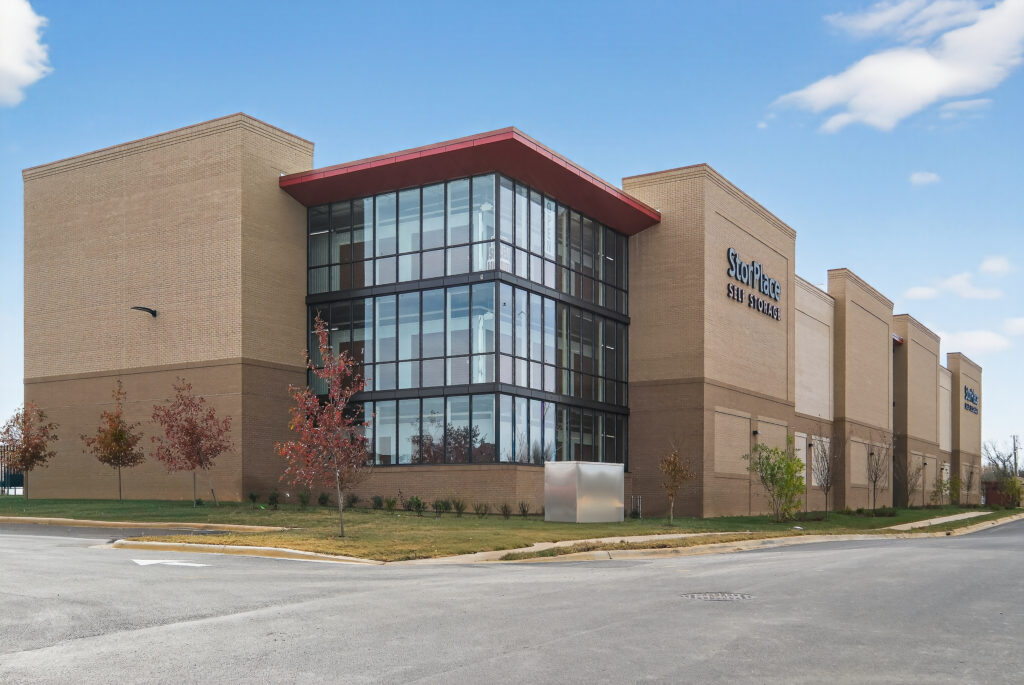 Modern tan brick StorPlace Self Storage facility with a multi-story glass corner entrance under a clear blue sky.
