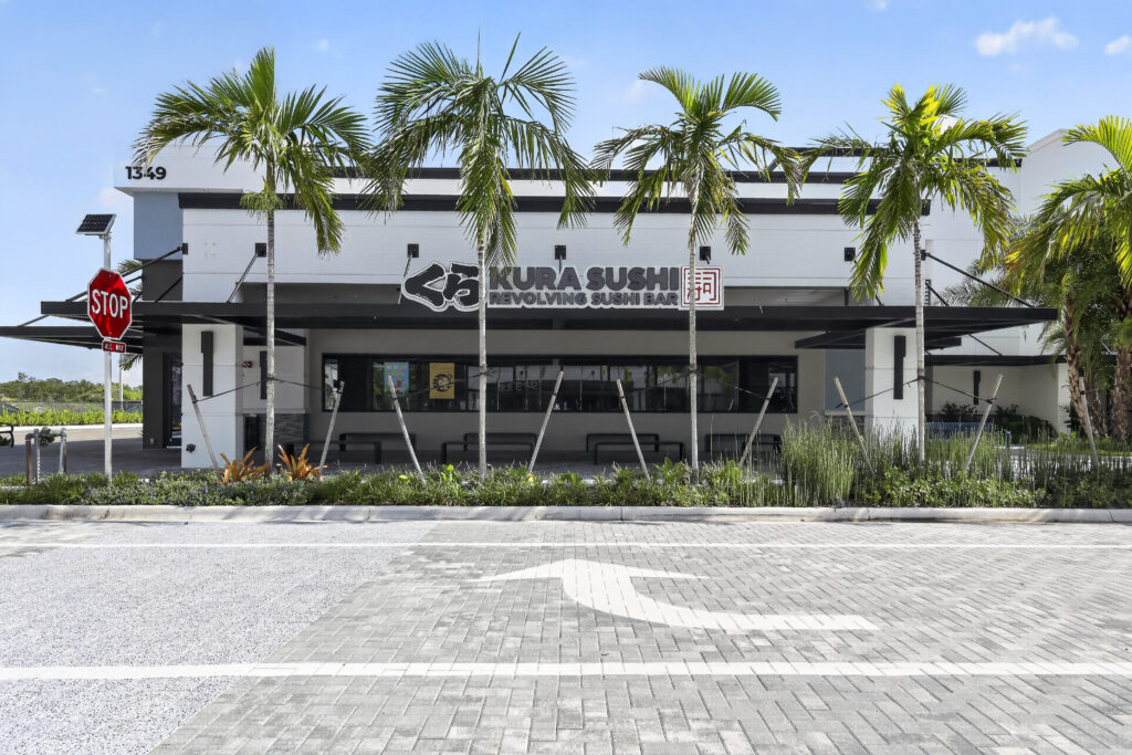 The exterior of a Kura Sushi restaurant building with palm trees in front and a stop sign on the left.