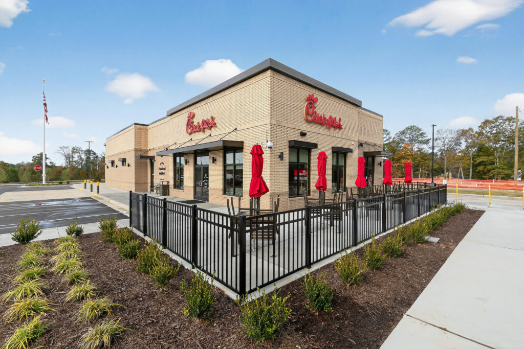Exterior view of a Chick-fil-A restaurant with outdoor patio seating under red umbrellas, surrounded by a black metal fence.