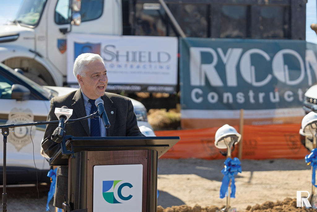 A man in a suit speaks at a podium in front of construction trucks and signs for Shield Contracting and Rycon Construction.