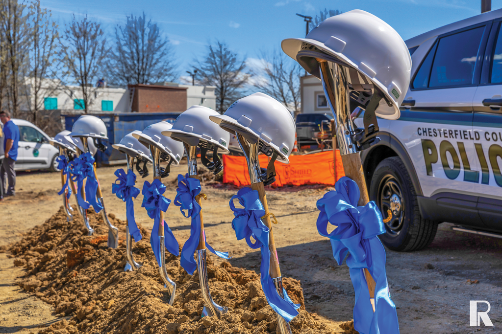A line of shovels with white hard hats perched on the handles, each decorated with a blue ribbon, stuck into the dirt at a groundbreaking site next to a police vehicle.