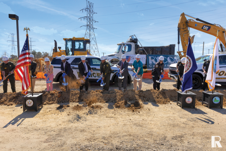 A group of officials wearing hard hats perform a ceremonial groundbreaking with shovels at a construction site.