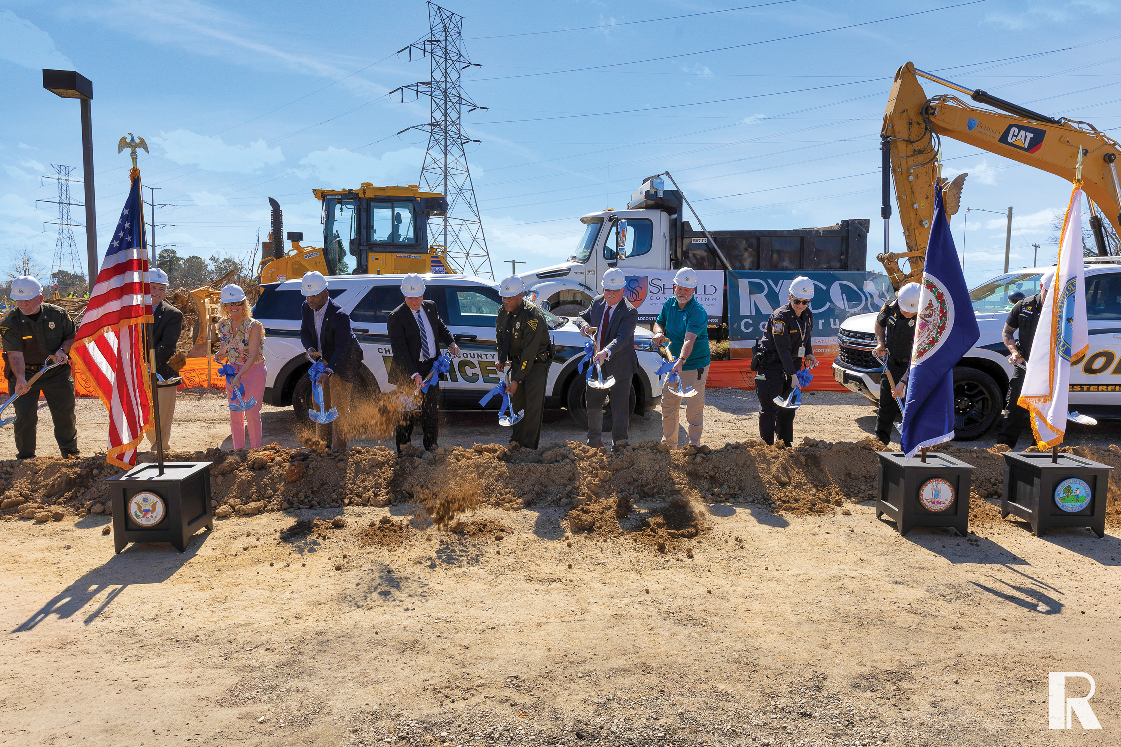 A group of officials wearing hard hats perform a ceremonial groundbreaking with shovels at a construction site.