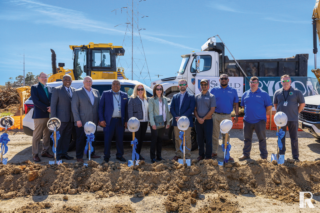A group of eleven people standing in a row on a construction site, each holding a ceremonial shovel with a blue bow. Construction vehicles are parked in the background.