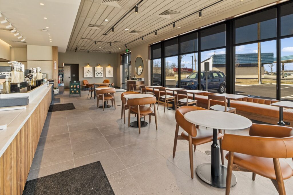 Interior of a modern coffee shop with a service counter on the left and seating consisting of small round tables and tan leather chairs.