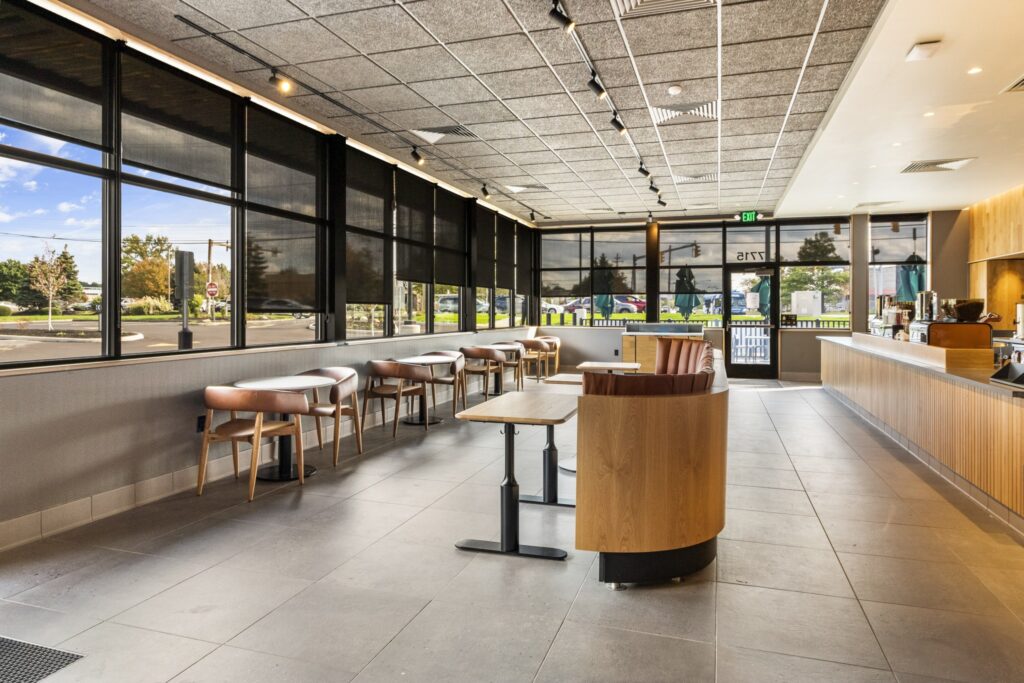 Interior of a cafe with tables and chairs, a curved booth, and a service counter along the wall, viewed through large windows looking out to a parking lot.