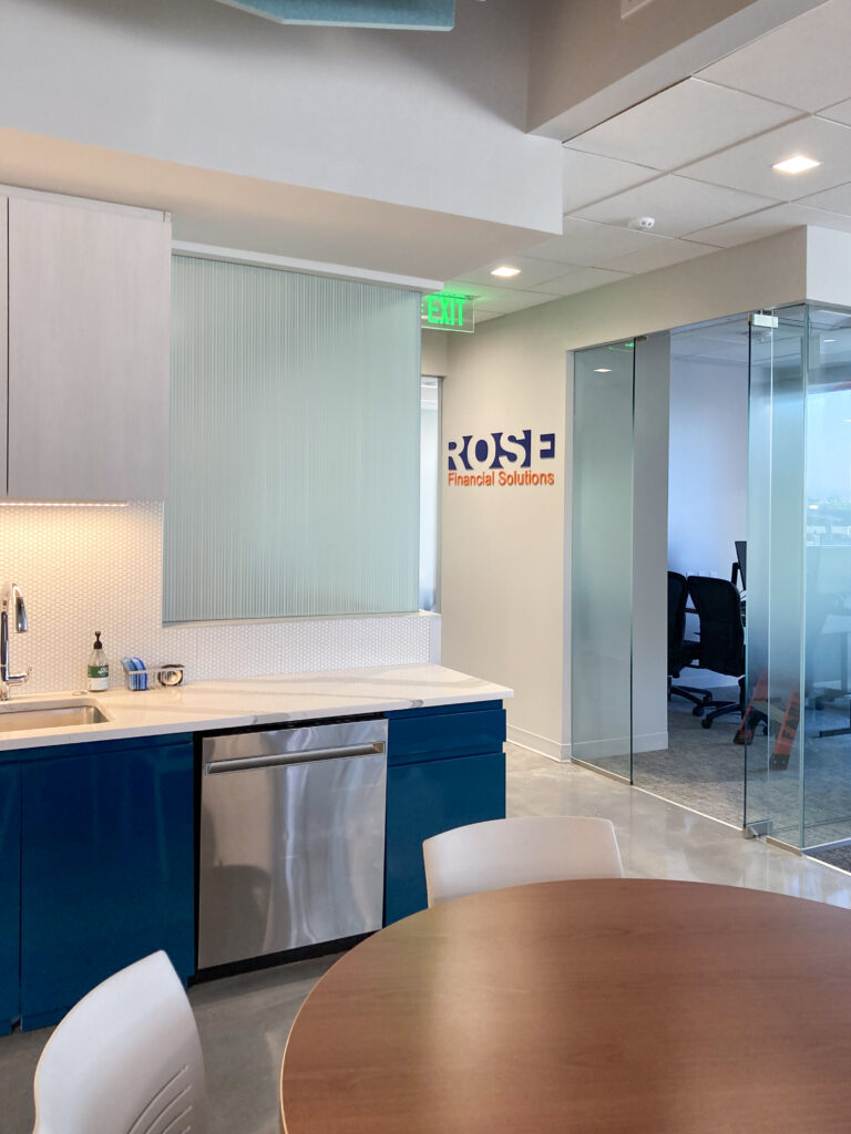 A modern office kitchenette with blue cabinets and a stainless steel dishwasher, with a “ROSE Financial Solutions” sign on the wall and a glass-enclosed conference room nearby.