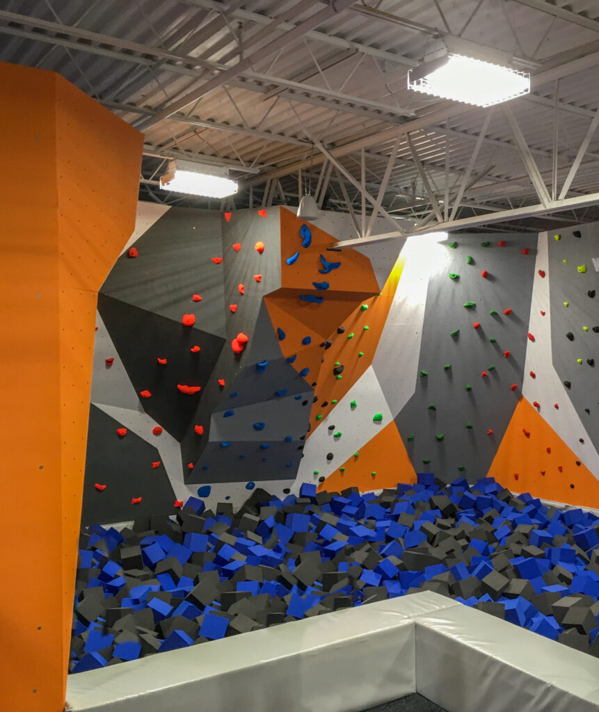 An indoor rock climbing wall with varied colored handholds above a pit filled with blue and black foam blocks.