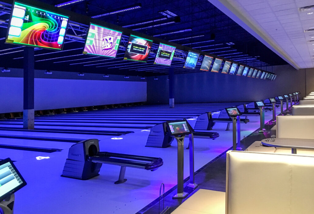Bowling alley with rows of lanes, ball returns, and overhead scoring monitors under blue mood lighting.