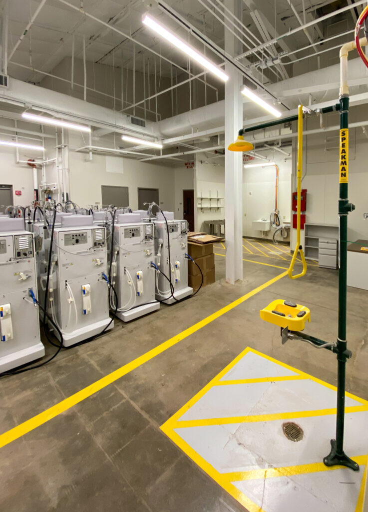 A row of medical dialysis machines inside a facility with a yellow emergency eye wash station in the foreground.