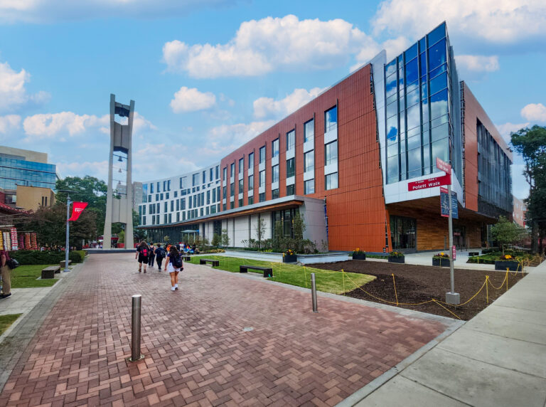 People walking along a brick path on a college campus next to a large, modern brick and glass building with a bell tower.