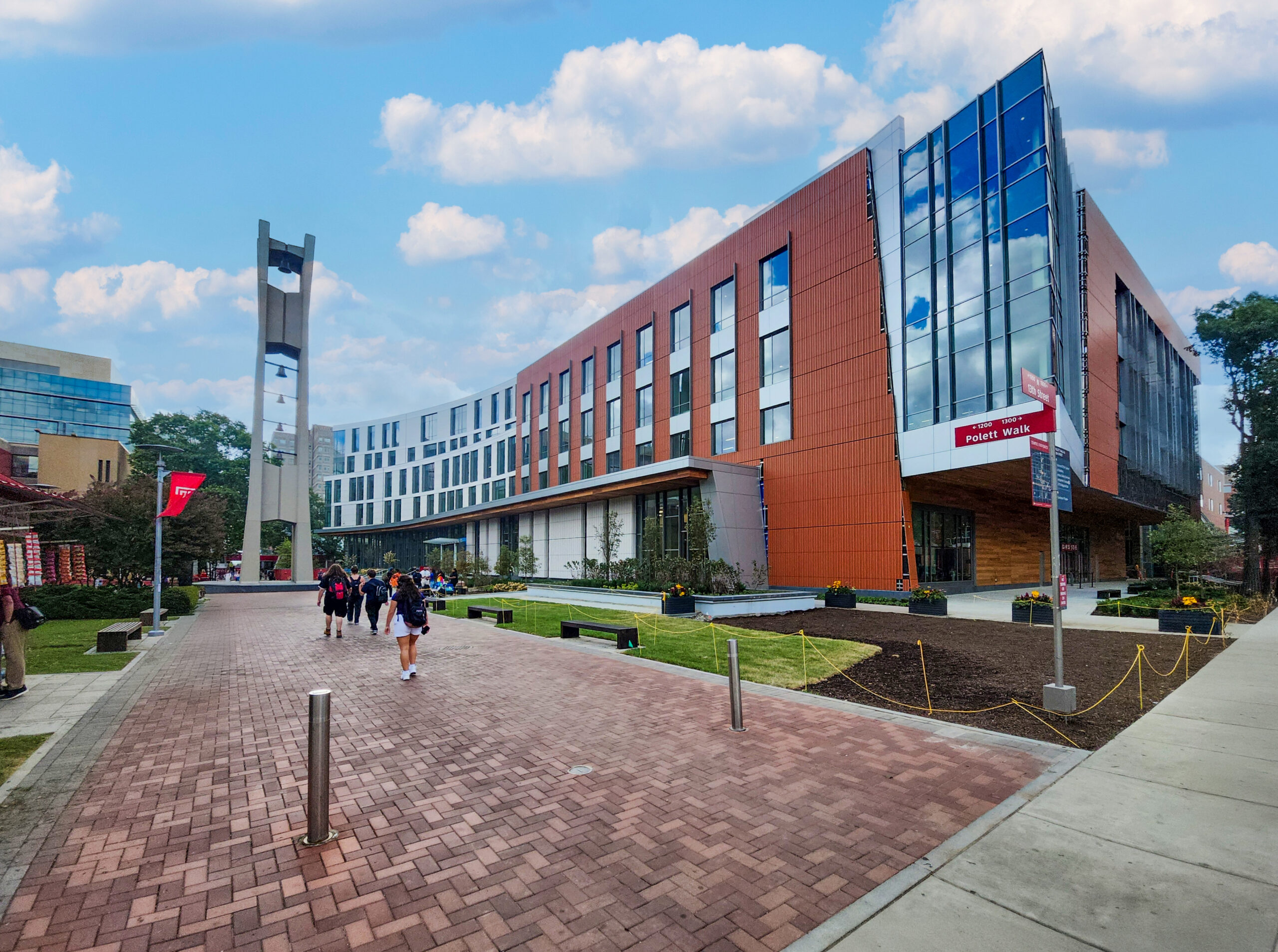 People walking along a brick path on a college campus next to a large, modern brick and glass building with a bell tower.