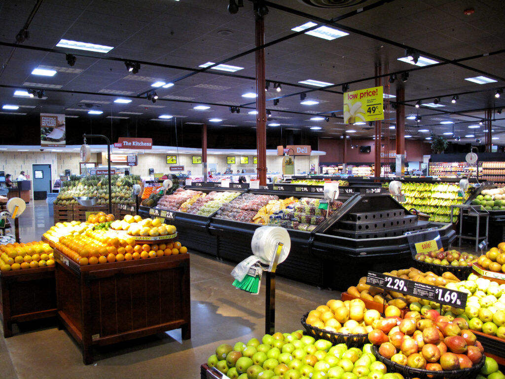 Produce section in a grocery store featuring bins of fresh citrus and apples with refrigerated displays of grapes and other fruit in the background.