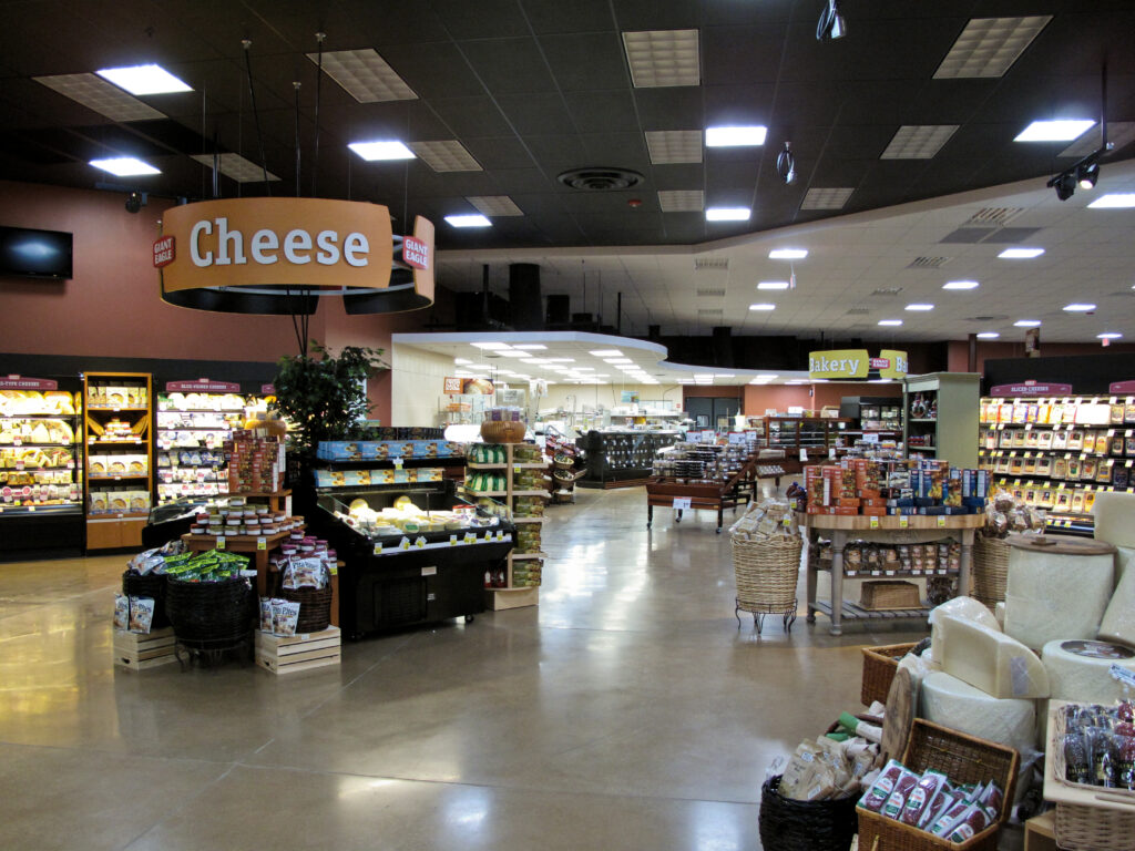 Inside a grocery store with a prominent “Cheese” sign hanging above a refrigerated display case, with bakery aisles visible in the background.