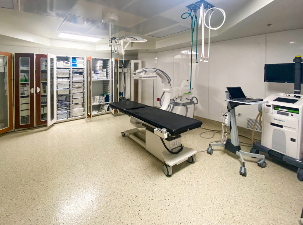 An examination room featuring a procedure table, a mobile C-arm imaging system, a medical workstation with a laptop, and cabinets filled with medical supplies.