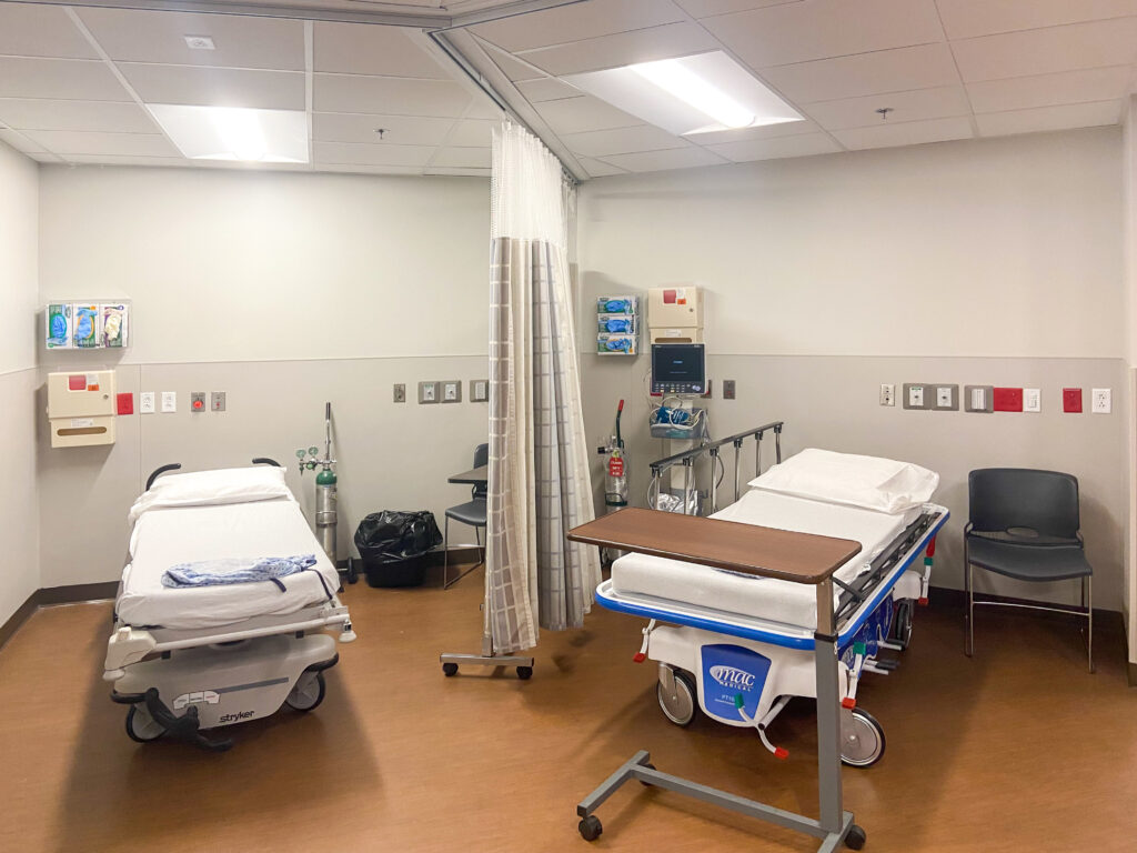 Hospital room with two patient stretchers, medical equipment, a privacy curtain, and chairs.
