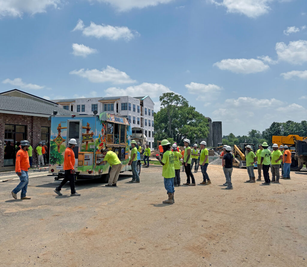 A group of construction workers in high-visibility vests and hard hats line up at a food truck on a job site.