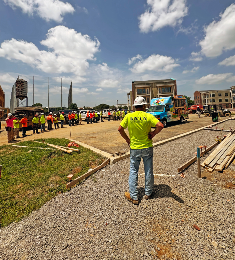 A construction worker in a neon yellow shirt stands on a gravel lot facing a line of workers and a parked ice cream truck.