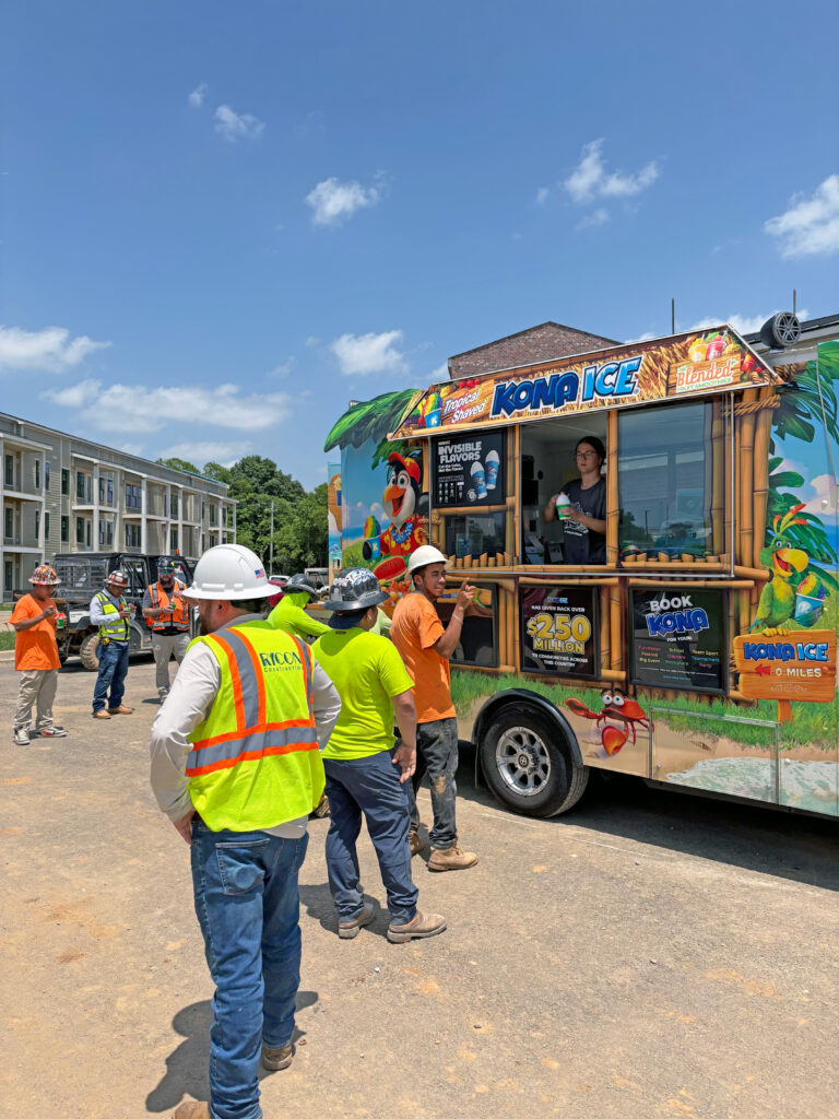 A group of construction workers in hard hats and high-visibility vests wait in line at a colorful Kona Ice truck.