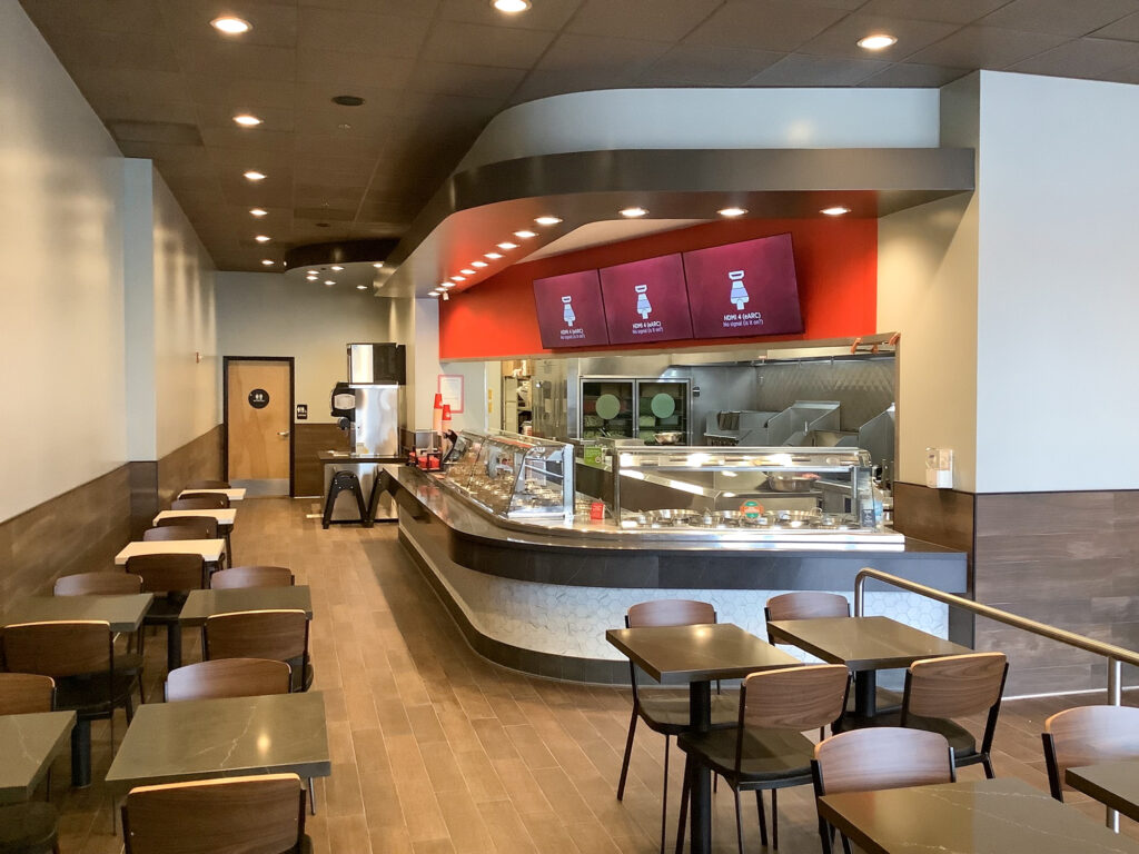 Interior of a fast-casual restaurant showing a food service counter with menu screens above and dining tables in the foreground.