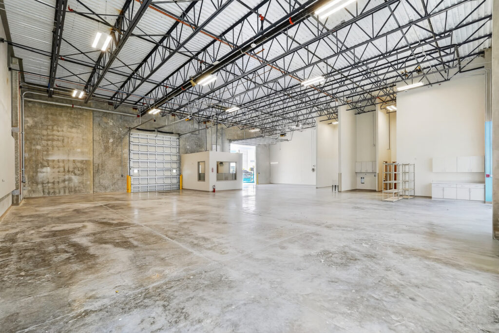 A large, empty warehouse interior with a concrete floor, high ceilings, an enclosed office area, and a roll-up bay door.