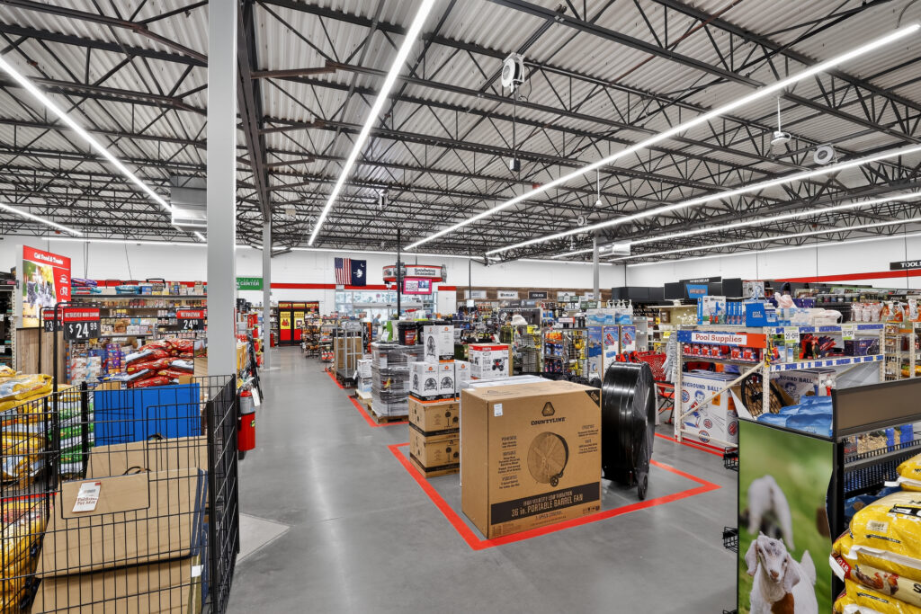 Interior view of a farm and home supply store with wide aisles, metal shelving units, and pallet displays of fans and other merchandise.
