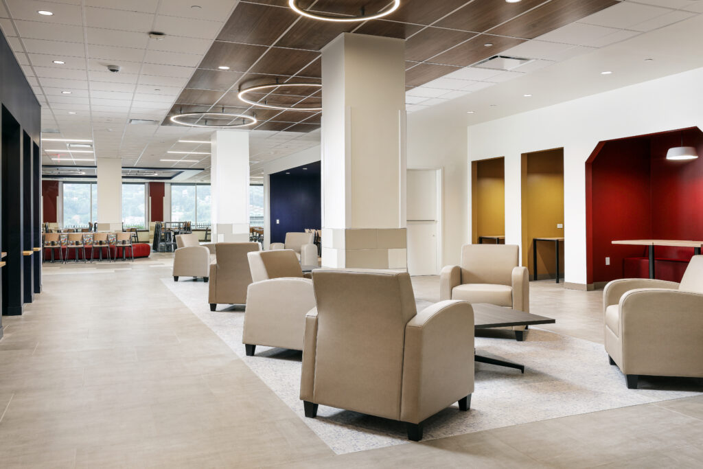 A large, open office lounge area featuring multiple beige armchairs arranged around low tables, with colorful recessed seating nooks along the back wall.