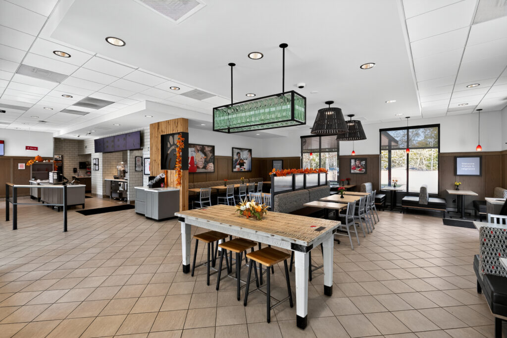 Interior of a restaurant dining area with a mix of tables, booths, and high-top seating, featuring neutral tile flooring and a service counter in the background.