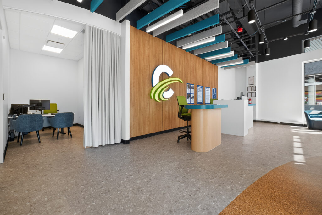 Modern office lobby featuring a reception desk, a wood-paneled wall with a large corporate logo, and an adjacent private office space behind a curtain.