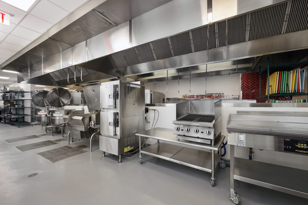 A commercial kitchen featuring stainless steel equipment, including a tilt skillet, a stacked convection oven, a gas grill, and a flat-top griddle under a large metal ventilation hood.